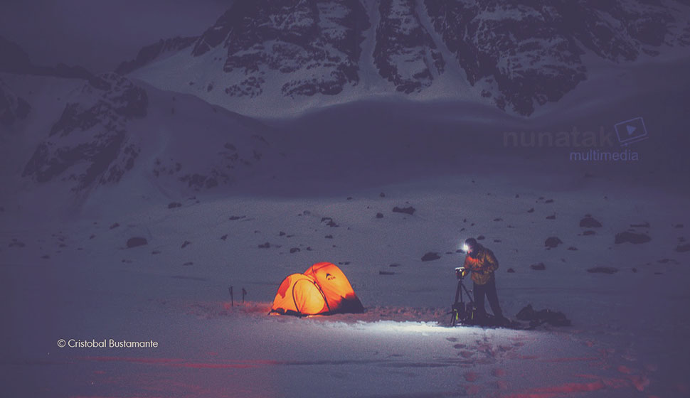 Campista con una tienda de campaña iluminada en la base de una montaña nevada por la noche, preparando equipo fotográfico, con el logo de Nunatak Multimedia en la esquina superior derecha.